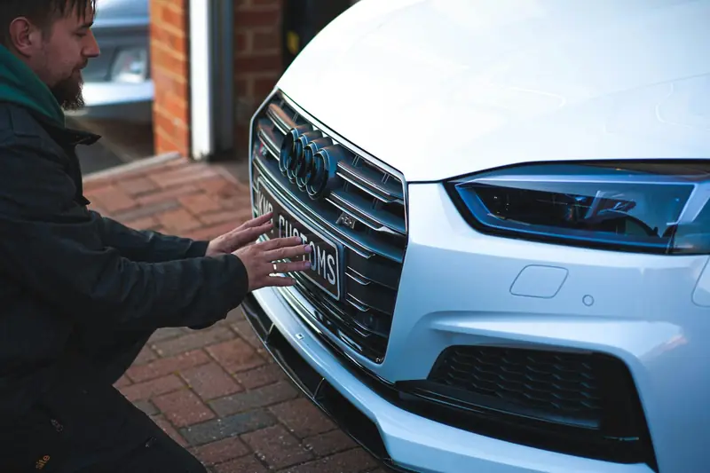 A mechanic kneeling at the front of a white car on a block-paved driveway