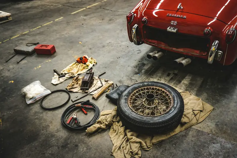 Tools, jump leads and a spare wheel laid out on a garage floor behind a red classic car