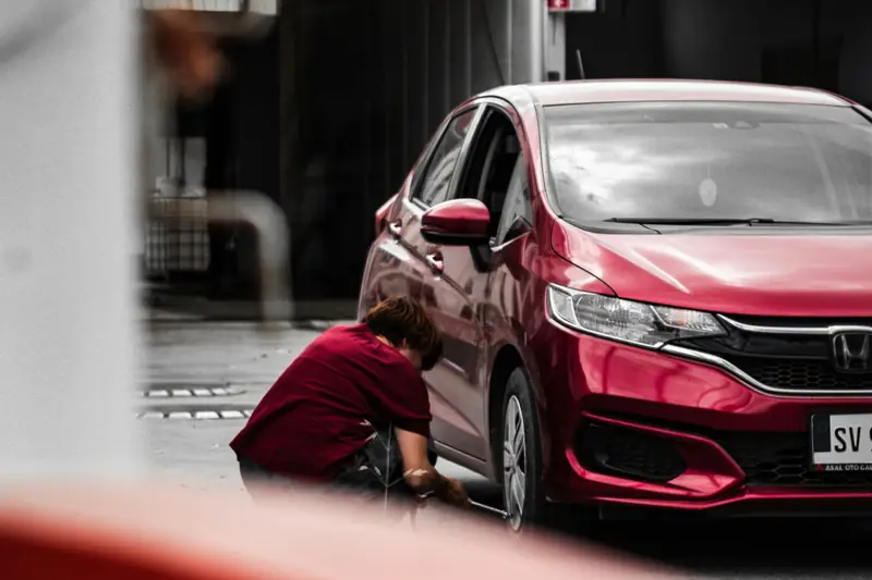 A mechanic crouched by the front wheel of a red hatchback